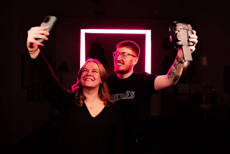 Joel och Mathilda står framför en lysande rosa led-fyrkant. mathilda tar en selfie med mobil och Joel tar en selfie med kamera